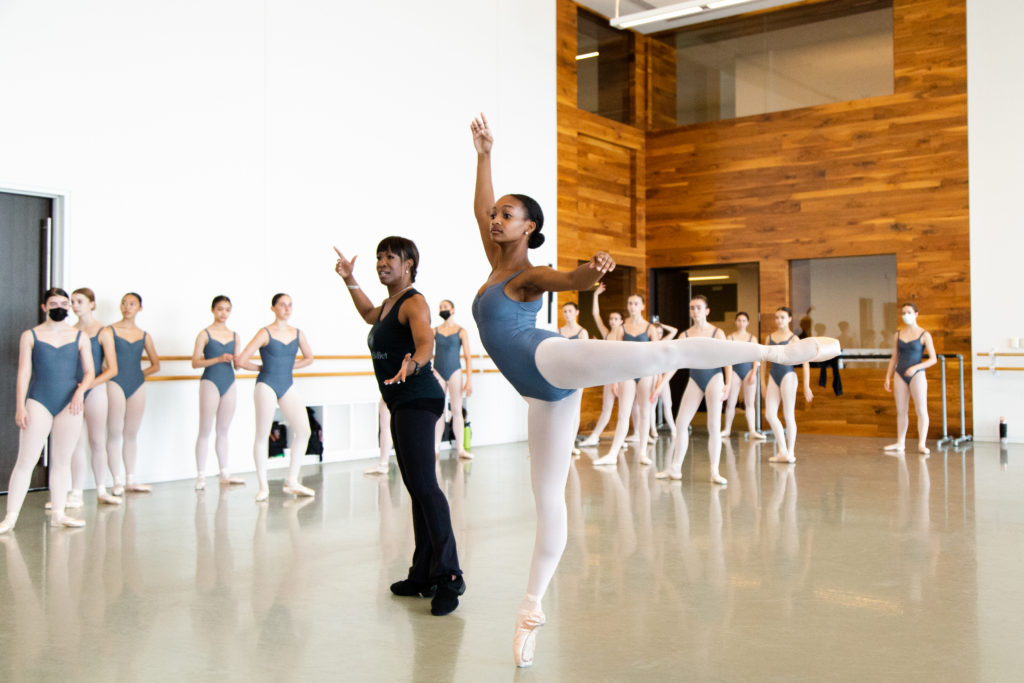 Lauren Anderson, dressed in a black tank top and yoga pants, coaches a Black teenage ballet student as she does a piqué arabesque on pointe. The rest of the class stands near the dance studio walls and watches. All of the girls wear gray leotards, pink tights and pink pointe shoes.