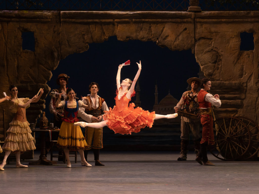 Skylar Brandt does a giant leap with her arms in high fifth position, looking up triumphantly and smiling joyously. She wears an orange-red, Spanish knee-length tutu with Spanish-style ruffles and holds a red fan in her upstage hand. Behind her, other dancers in Spanish-style costumes clap and smile, cheering her on. The set shows a tavern scene.