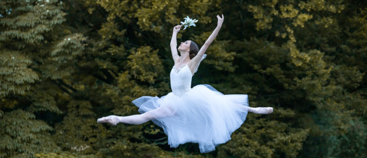During an outdoor performance at dusk, Skylar Brandt does a giant saut de chat with her arms opening out of high fifth position. She holds a white lily in her upstage hand and looks toward the sky forlornly. She wears a long, white Romantic-style tutu, pink tights and pointe shoes. She dances on a dark marley stage floor, and in front of green evergreen trees.