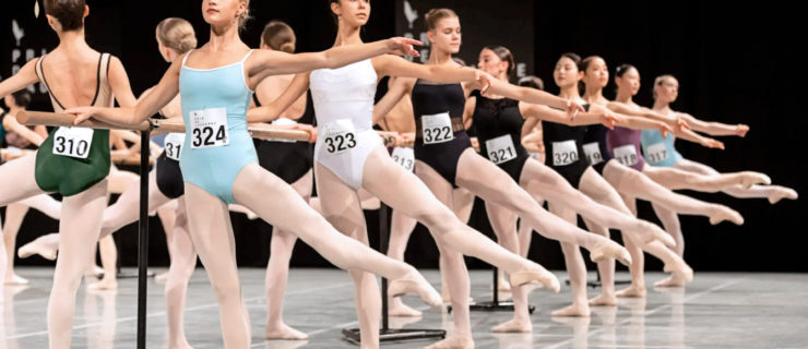 On a stage with a light grey floor and black backdrop, a long row of young international female ballet dancers execute a dégagé exercise at barre. They wear solid colored leotards with individual numbers pinned to the front, pink tights and ballet shoes.