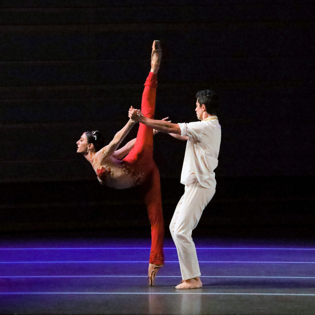 Tara Ghassemieh, wearing a sheer, red, one-piece costume with flesh-toned mesh fabric on the sleeves and torso, is in an assisted penché, supported by her husband, Vitor Luiz.