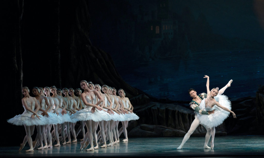Skylar Brandt and Herman Cornejo perform a somber pas de deux in Swan Lake. Brandt, in a white classical tutu and tiara, poses in a high attitude derriere, her eyes downward, as Cornejo, in white tights and a green medieval tunic, holds her tenderly by the waist. To their right, two lines of female dancers in similar white classical tutus pose with their left foot back in B plus and their hands crossed in front of them, their eyes looking sadly down towards their right.