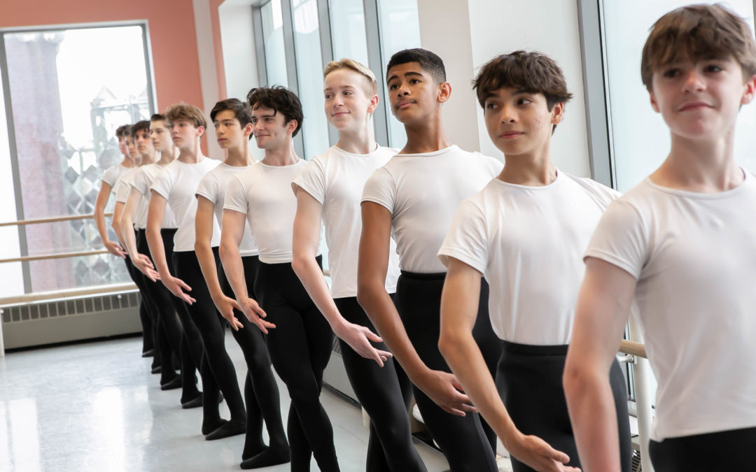 Eleven teenage male ballet students stand at the barre against a wall of windows in a dance studio. They all pose in fifth position with their left hand on the barre and their right arm en haut, their heads looking out towards their right. They all wear white T-shirts tucked into black tights, as well as black ballet slippers.