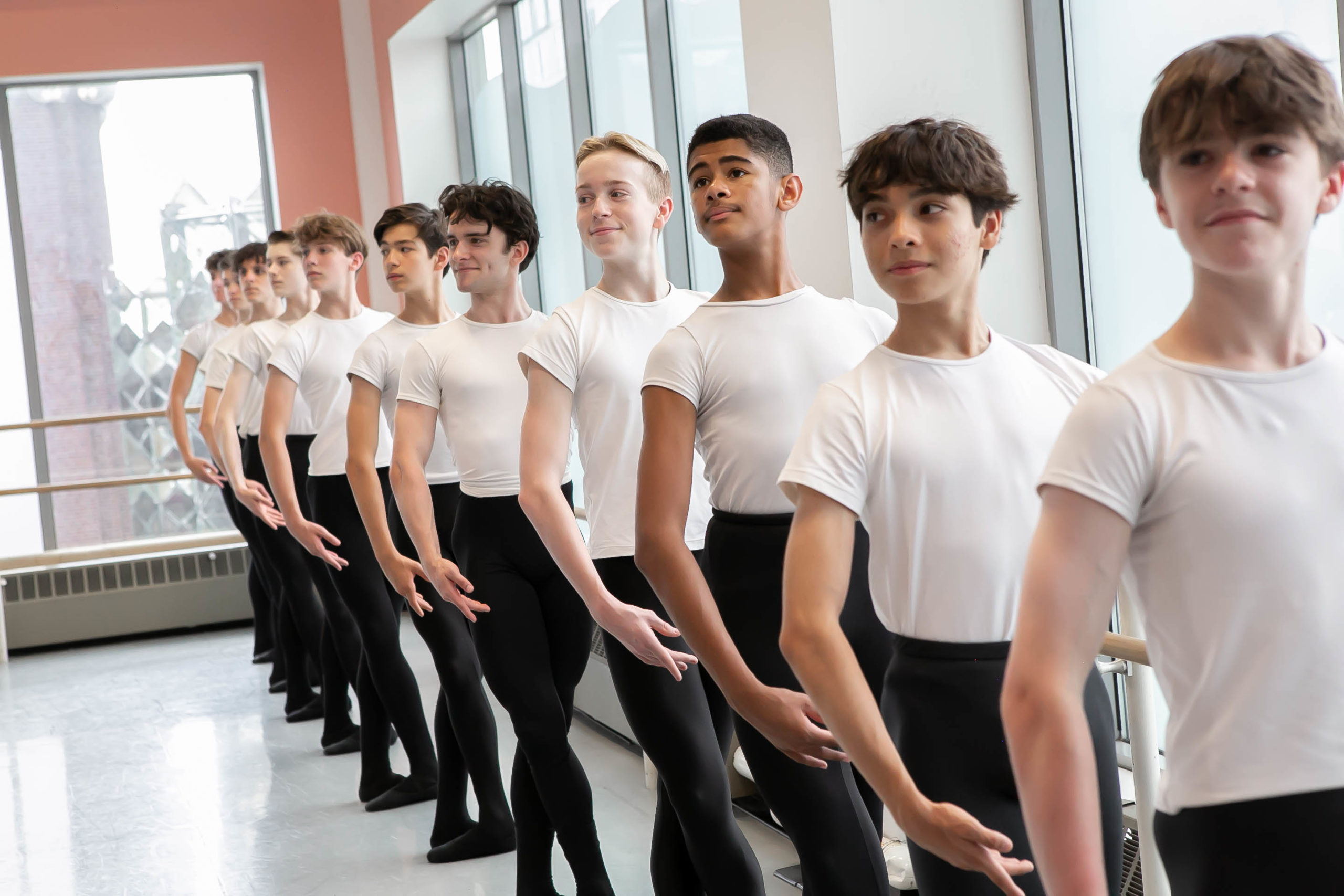 Eleven teenage male ballet students stand at the barre against a wall of windows in a dance studio. They all pose in fifth position with their left hand on the barre and their right arm en haut, their heads looking out towards their right. They all wear white T-shirts tucked into black tights, as well as black ballet slippers.