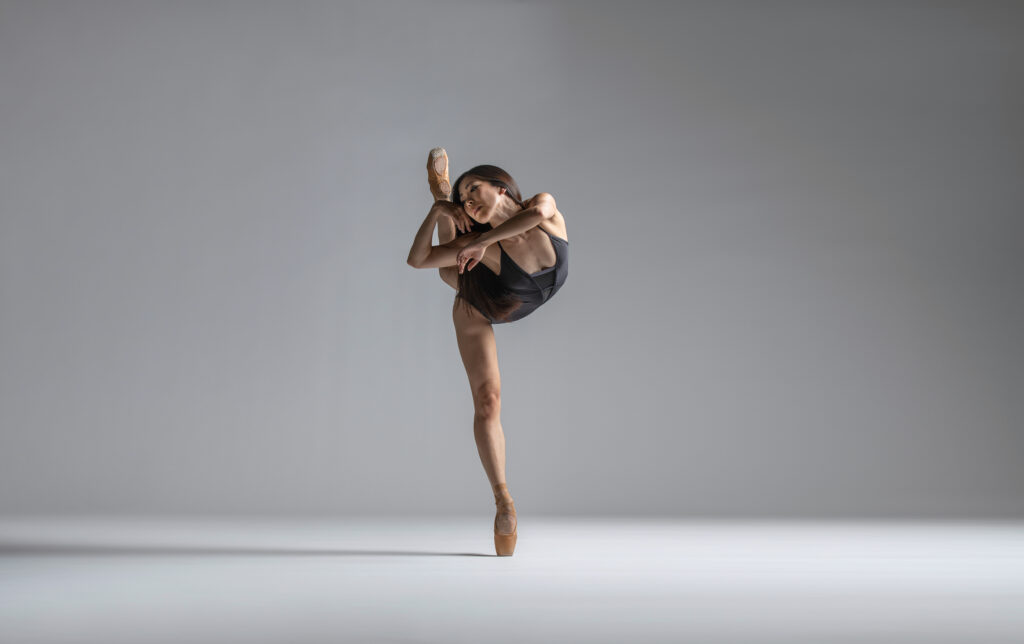 Andrea Yorita stands on her right foot on pointe and kicks her left leg back behind her, in parallel with a bent knee. She arches back and to her right with her upper body, curling her right arm in so that her hand touches her cheek and reaching her bent left arm across her chest. She wears a black leotard and tan pointe shoes.
