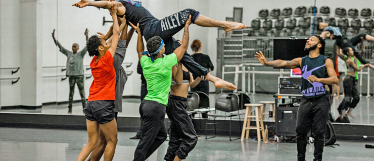 In a large dance studio, a group of 2 male and 2 female dancers lift a male dancer above their heads as he lies back, arms extended and legs gently bent. A male choreographer watches and gives direction from the right. They all wear various active and dance-wear clothing in shades of black, lime green and red.