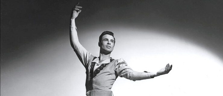 In this black and white photo, a male danseur, John White, is shown from the waist up, lifting his curved right arm up and his left arm out to the side, palm, up. He wears a costume, a light-colored jacket with circular embellishments on the lapels and a belt at the waist, and a collared shirt underneath. He looks over his left shoulder slightly, eyes raised slightly up, and with a small, open-mouthed smile.