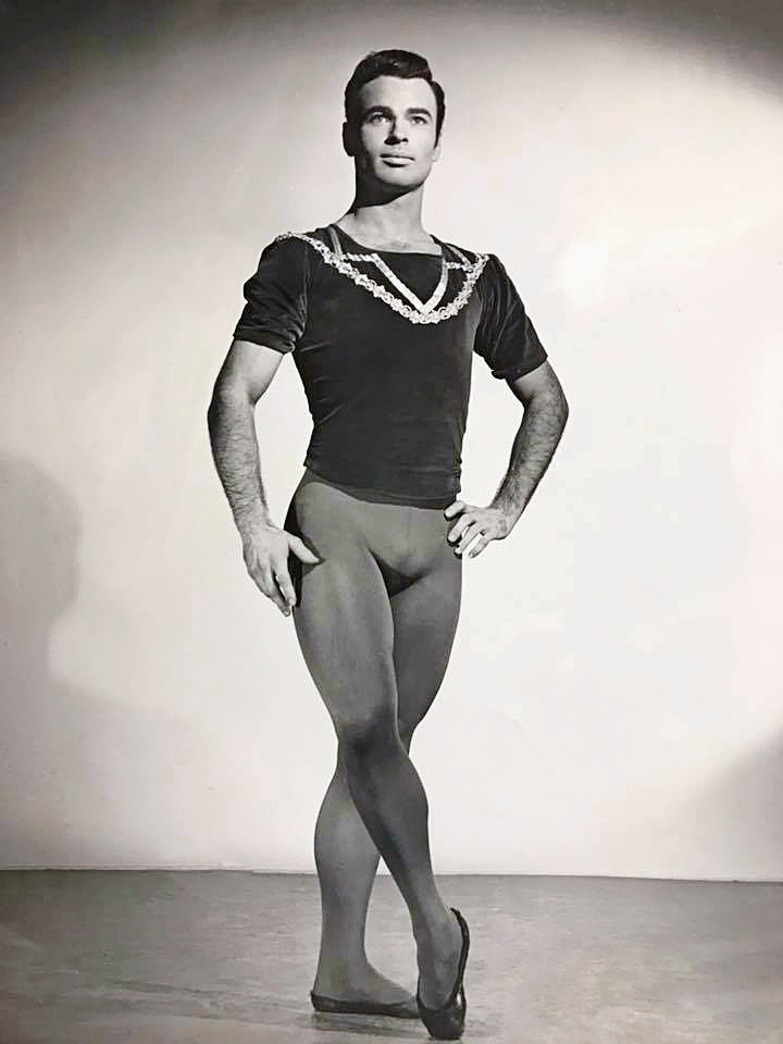 IN this black and white photo, John White poses for the camera, standing casually with his right foot crossed in front and popped up onto full pointe. His left hand grazes his left hip, and his right arm rests gracefully at his side. He wears a dark, short-sleeved top with shiny embellishments near the neckline, dark tights and dark ballet slippers. He looks off into the distance with a peaceful countenance.