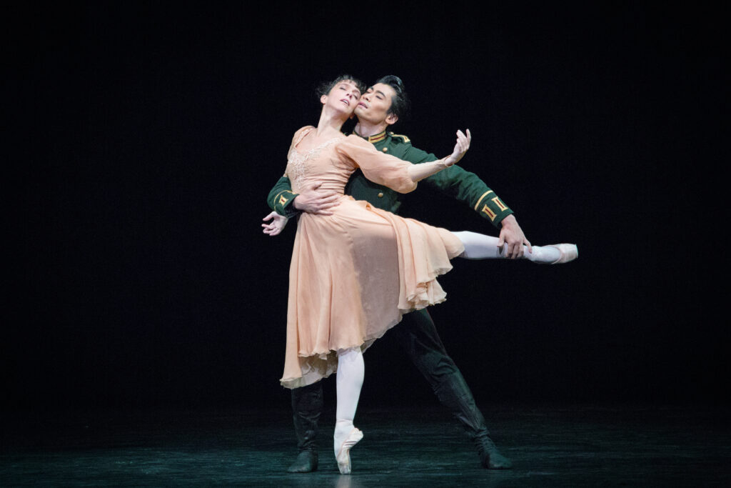 On a dark stage, two dancers perform a pas de deux together. The woman wears a light peach flowy dress and stands on pointe in a low arabesque, arched back against her partner as she extends her left arm back as if to cradle his head. The man, in a dark green military top and black tights, stands behind her in tendu as he supports her and gently holds her arabesque leg.