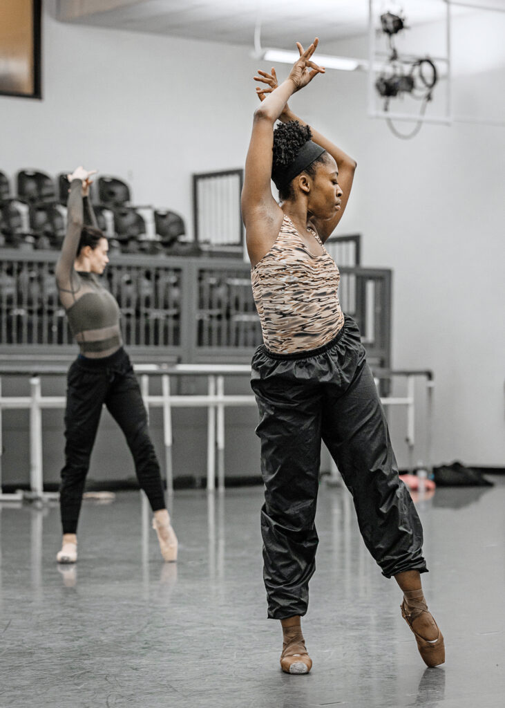 In a large dance studio, two female dancers pose with their arms bent over their heads, and looking to the left. They shift their weight to their right leg, left legs parallel and pointed. They wear leotards, black athletic pants and pointe shoes.