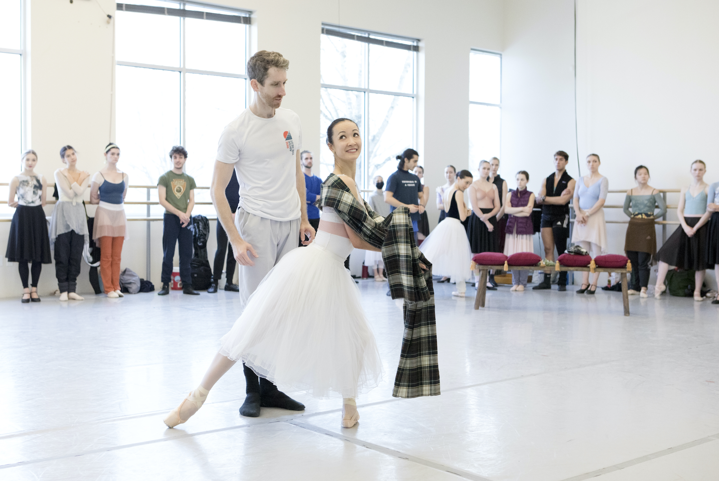 Xuan Cheng and Bria Simcoe rehearse a scene from the ballet La Sylphide in a large dance studio with large windows. Cheng, in a long white tutu, lunges in front of Simcoe with her right foot in tendu derriere and wraps a plaid tartan shawl around her shoulders. She looks over her right shoulder and up at Simcoe coyly as he stands behind her. He wears a white T-shirt, gray sweatpants and black socks and ballet slippers, and looks at Cheng intently. Towards the back of the studio, a large group of dancers in practice clothes stand and watch them rehearse.