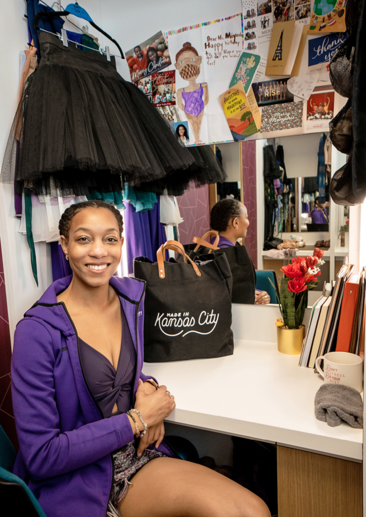 McGriff is smiling at the camera as she sits in her dressing room in front of a mirror. She is wearing a purple warm-up jacket with her name embroidered on the left side. She is surrounded by her dance bag, mug, flowers, books, a black tutu, pointe shoes, other dance wear, and cards and drawings.