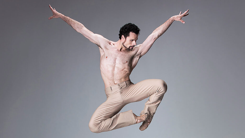 In front of a light gray background, a male dancer in nude jazz pants and ballet shoes jumps in a stylized pas de chat position with his arms extended in a wide "v" overhead. He looks down on the right diagonal.