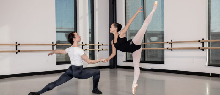 A male in a deep lunge holds her partner's hand as she does a piqué arabesque on pointe.