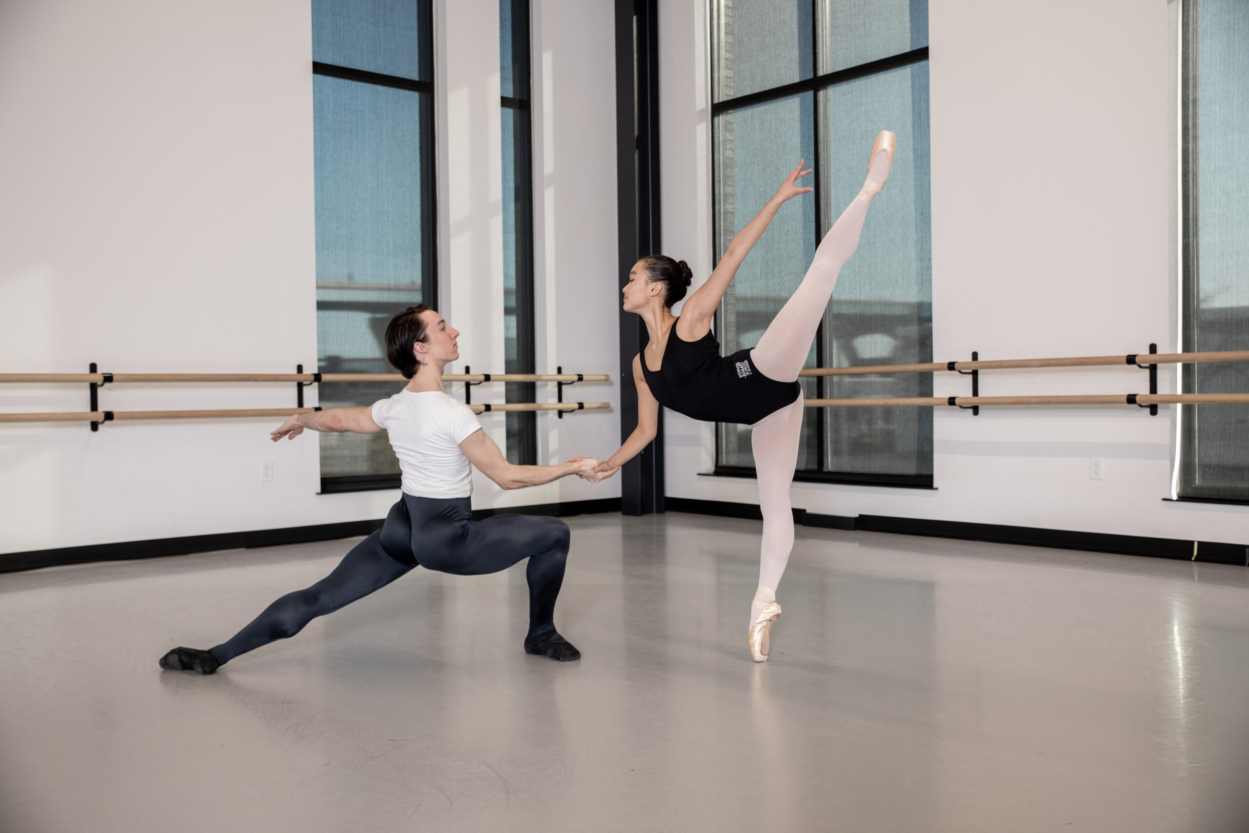 A male in a deep lunge holds her partner's hand as she does a piqué arabesque on pointe.