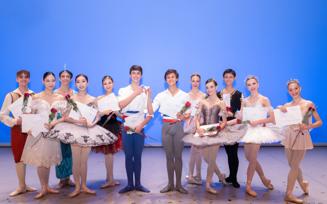 Onstage in front of a light blue backdrop, a group of 12 male and female dancers pose together and smile, wearing various ballet costumes and holding roses and awards. There is confetti at their feet.
