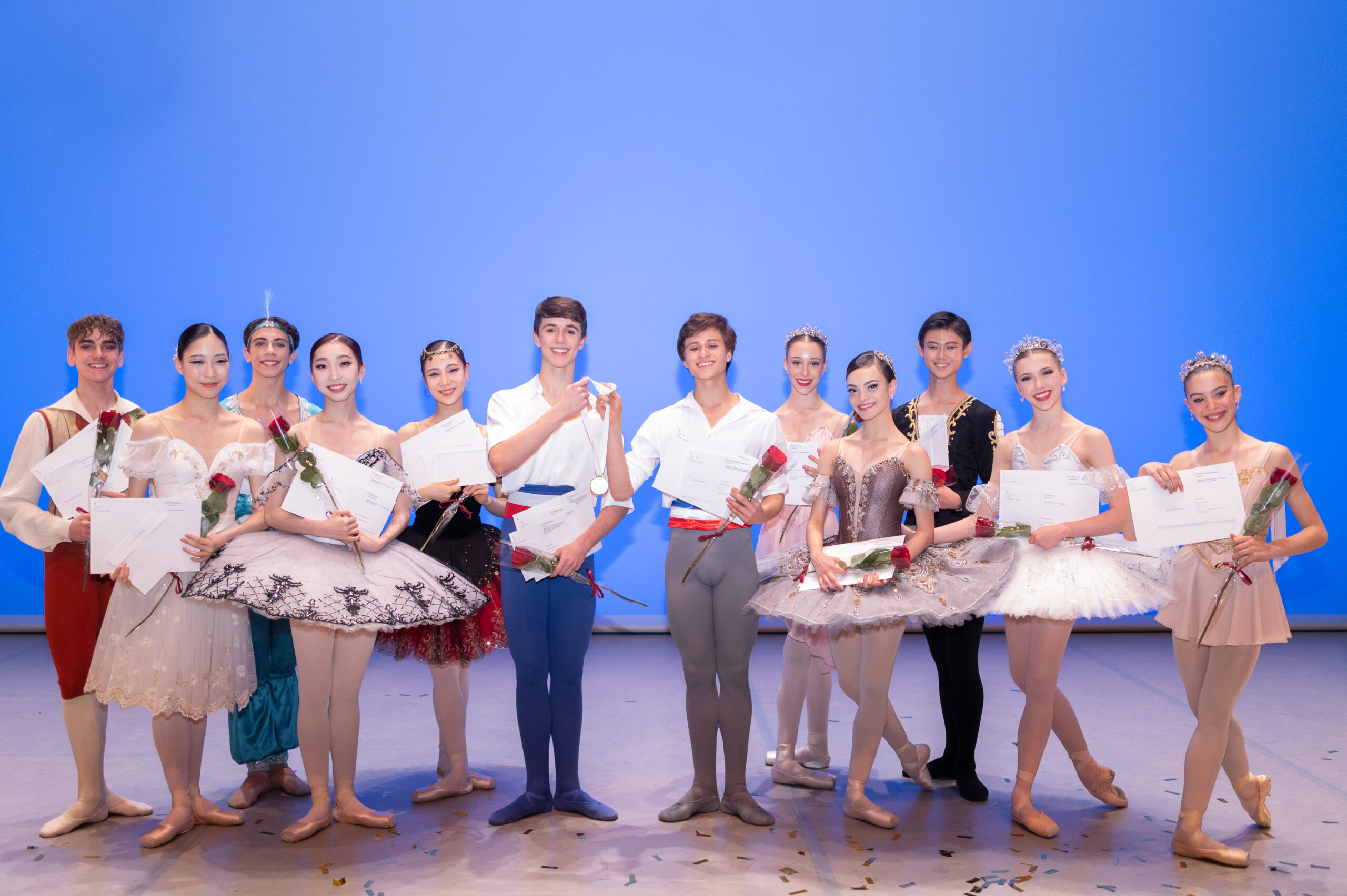 Onstage in front of a light blue backdrop, a group of 12 male and female dancers pose together and smile, wearing various ballet costumes and holding roses and awards. There is confetti at their feet.