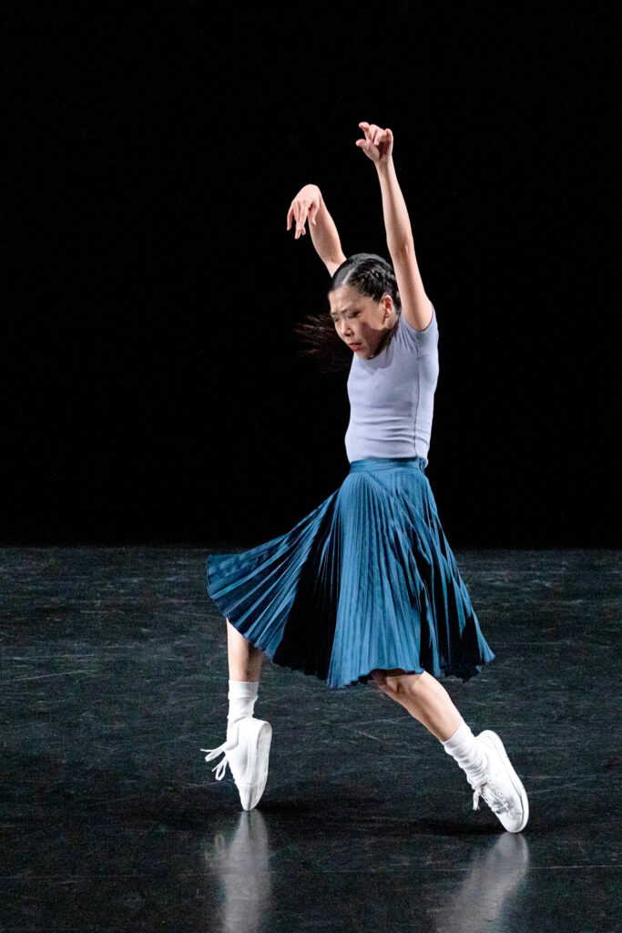 Andrea Yorita performs onstage in front of a black backdrop. She stands upright on the toe of her sneakers as if on pointe, and reaches both arms up as she looks towards the floor. She wears a pleated blue skirt, a gray short-sleeved top, white socks and white sneakers.