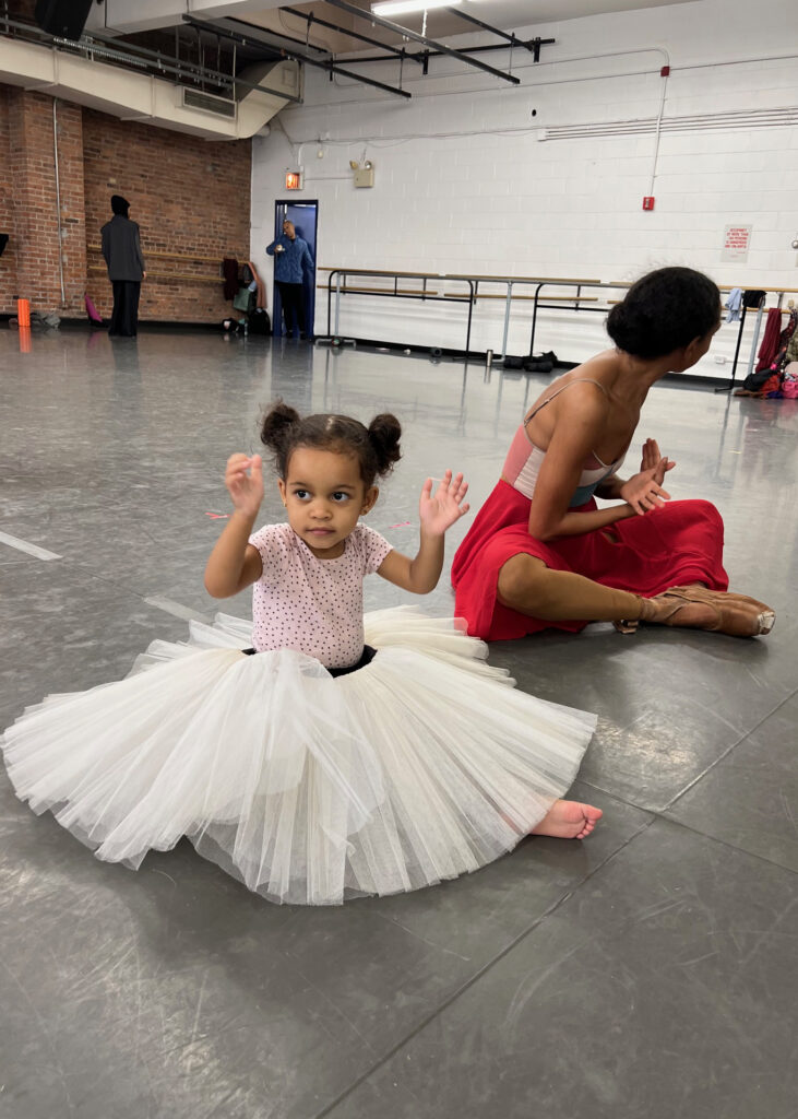 A little girl in a pink shirt and wearing an adult practice tutu sits on the floor of a dance studio. To her left sits a professional dancer in a long red skirt, brown pointe shoes and a pink and white leotard.