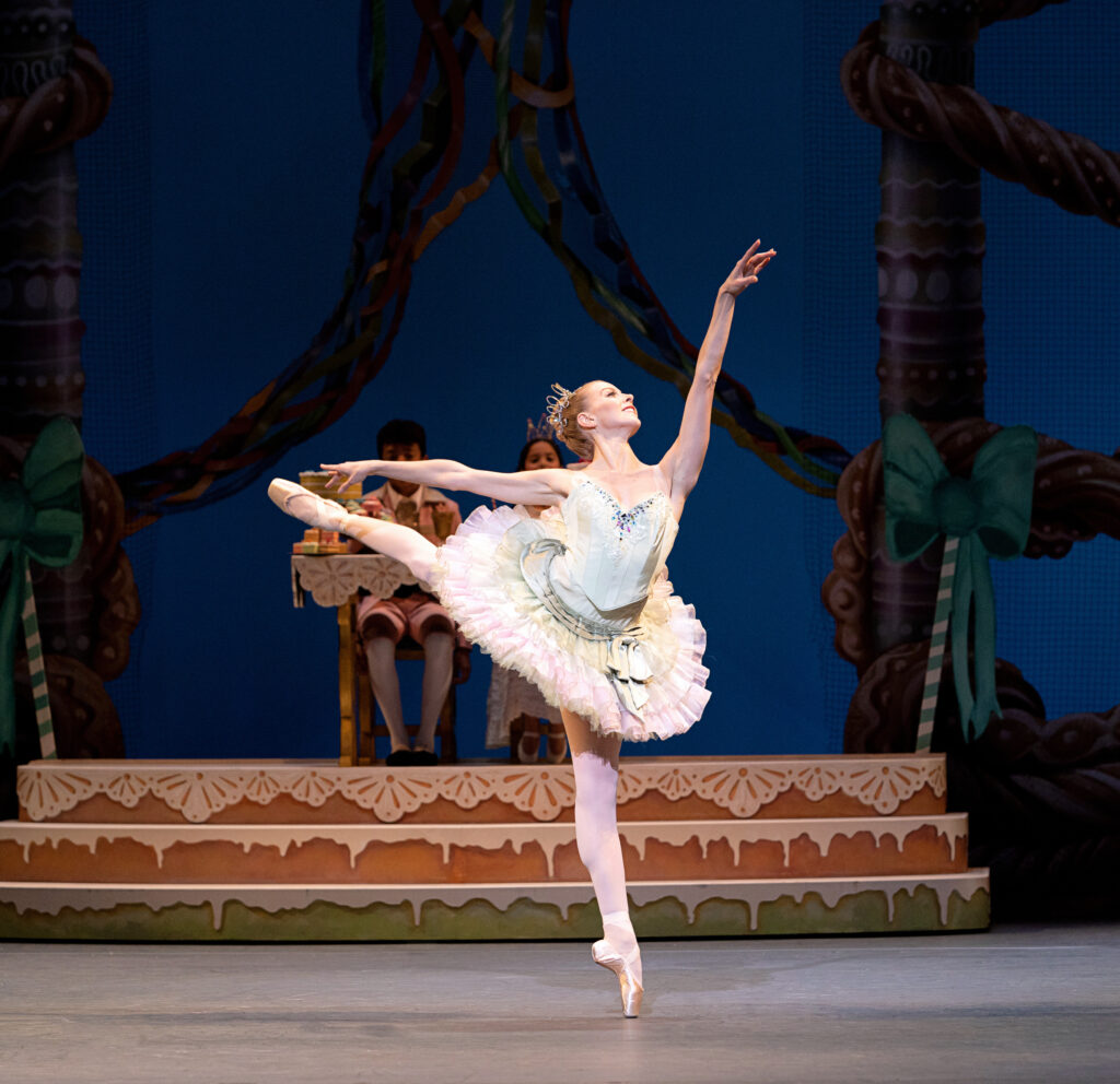 On an ornately decorated stage, a female ballerina in a light green and pink pancake tutu performs a pique arabesque. She radiates joy and confidence.