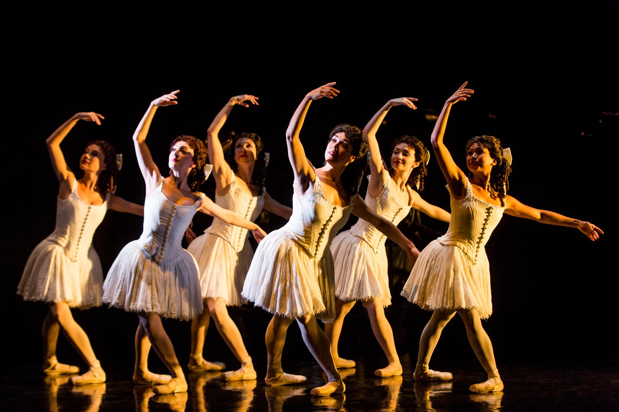 A crosp of six female ballet dancers stand in two staggered lines and pose in a croise fourth position plié with their right leg in front. They hold their arms in third position, looking underneath their raised right arm. All of the women wear knee-length tutus and corseted bodices, tights and pointe shoes, and wear their hair in curls, secured back with a large bow.