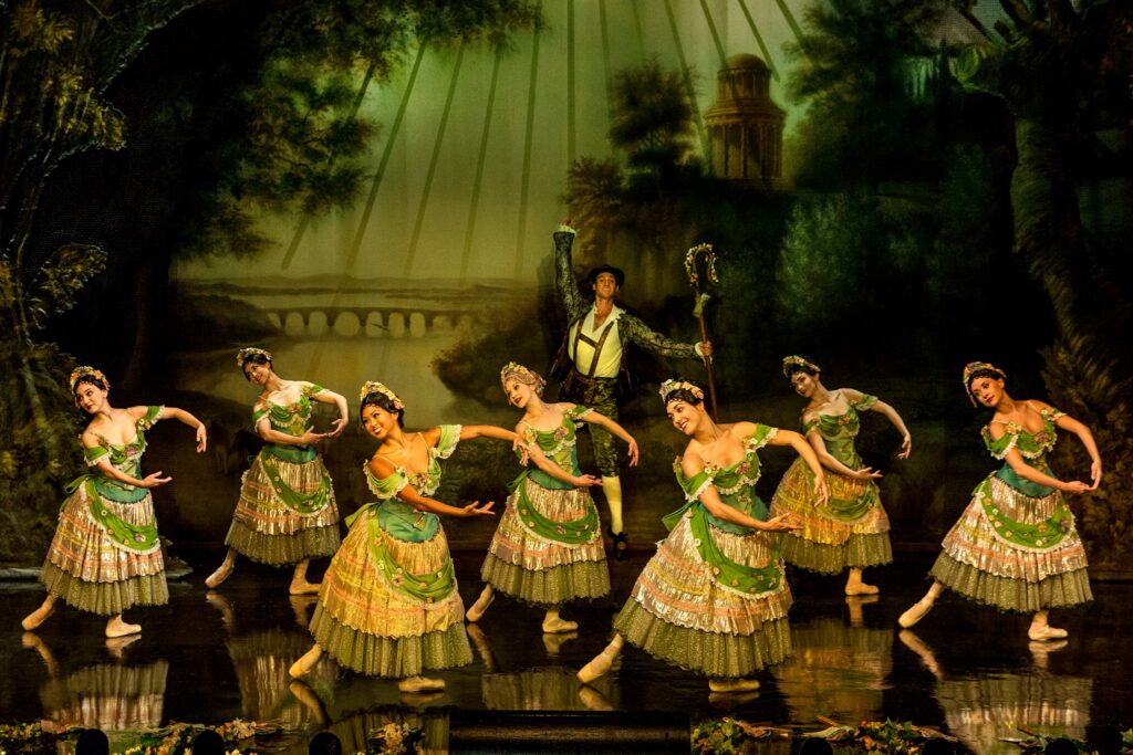 A corps of eight female dancers in long, ornate green dresses with floral headpieces pose in three staggered lines onstage. Wearing pointe shoes, they stand in a tendu derriere in effacé with their right legs back, and their arms curved into a first position and swinging out slightly to their left. They all look over their right shoulders with large smiles. A male dancer in lederhosen and a holding a shepherd's staff dances behind them, jumping up with his right arm above his head.