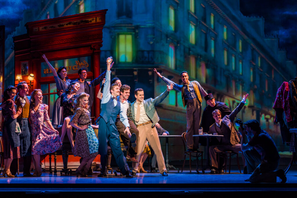 During a performance of An American in Paris, the ensemble gathers in a large group to pose for a picture and facing a photographer downstage. Many of them hold their arms up triumphantly in the air, and wear costumes reflecting the 1940s.