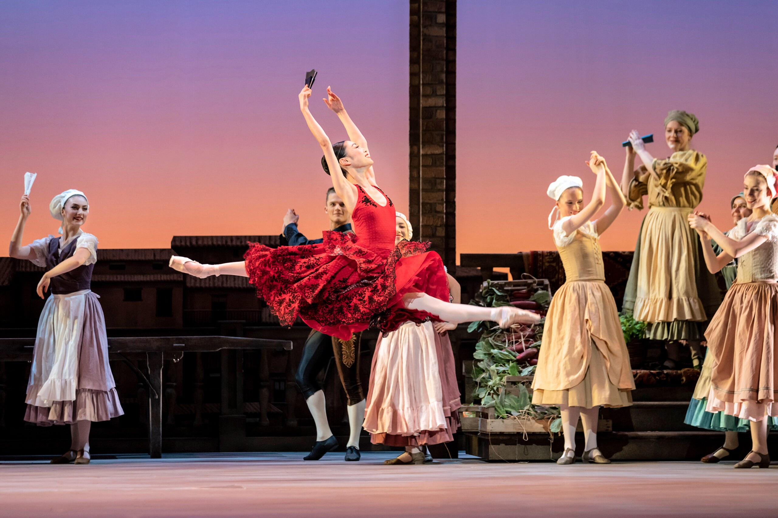 A ballerina in a red knee-length, Spanish style tutu, pink tights and pointe shoes does a saut de chat with her arms in high fifth. She holds a black fan in her right hand and looks up triumphantly as she jumps. Behind her onstage, six dancers wearing peasant costumes clap their hands up hig as they watch her.