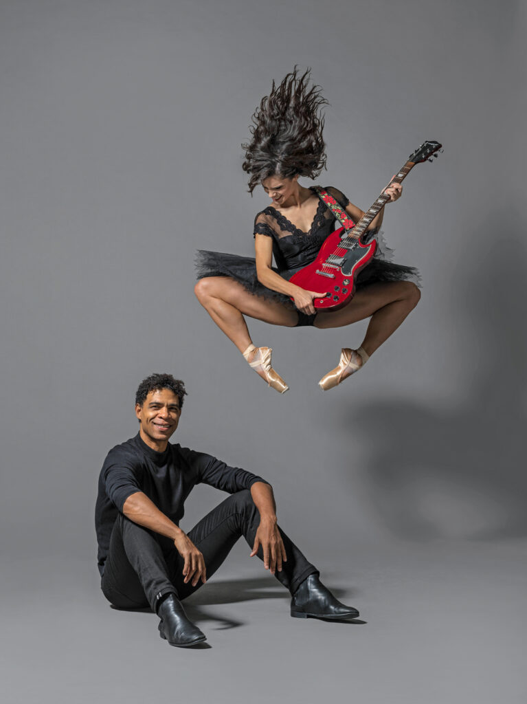 Carlos Acosta, wearing all black, sits on the floor with his knees propped up, his hands resting casually over them, and smiles twoards the camera. Behind him, Sofia Liñares jumps up, tucking her legs under her, and holds a red electric guitar, her long hair flying and her smiling face looking downward. She wears a black leotard with lace trim, a black practice tutu and pointe shoes. They pose in front of a gray backdrop.