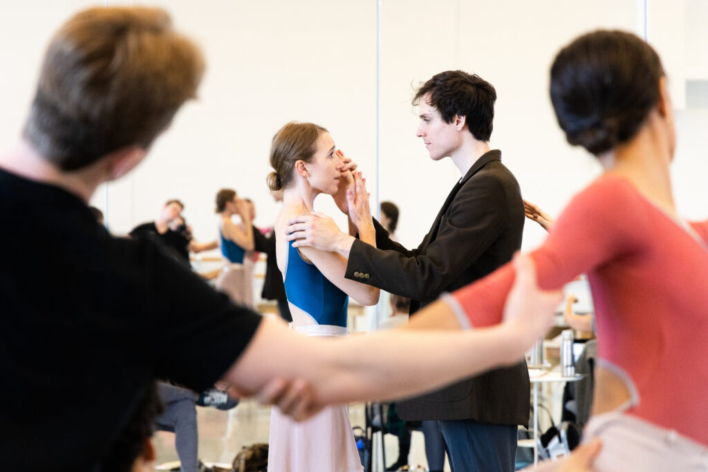 A male and a female dancer rehearse together in a bright studio, holding each other gently at a slight distance, as corps de ballet members link arms and dance in a circle around them.