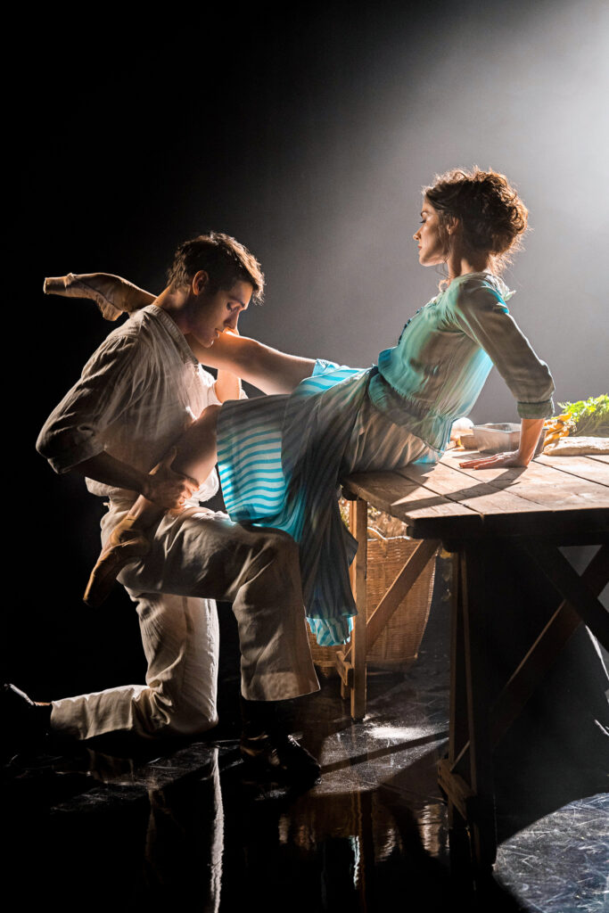 Cassandra Trenary, in a long light blue and white striped house dress, and Daniel Camargo, in a white button down and beige trousers, pose together sensually as Tita and Pedro in "Like Water for Chocolate." Trenary sits on a wooden table facing Camargo, leaning back on her hands slightly as she lifts her legs around him gently. Camargo kneels, her right leg on his shoulder as he holds her left calf and looks down at her leg passionately.