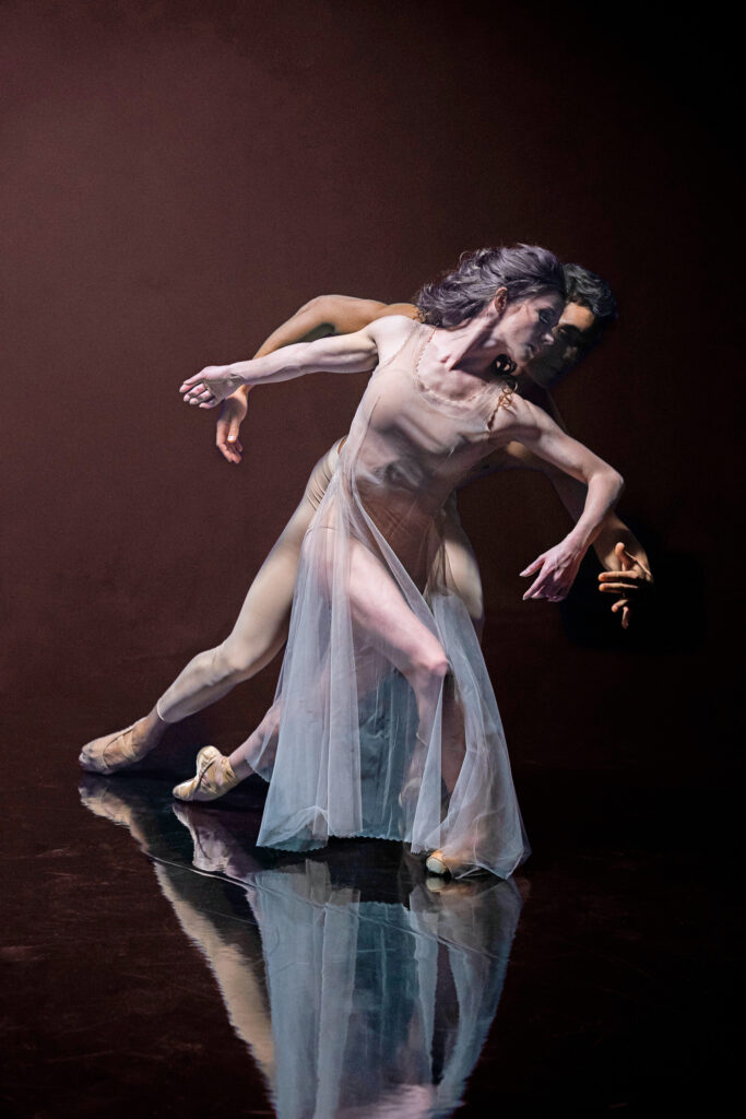 In front of a dark rust background, Cassandra Trenary and Daniel Camargo dance together as Tita and Pedro in "Like Water for Chocolate." Trenary wears a nude-colored dress with a sheer, wispy skirt that's slit on the side, and she lunges with her back to Camargo. Camargo wears nude tights and lunges with her, chest toward her back. Their arms trace each other with longing.