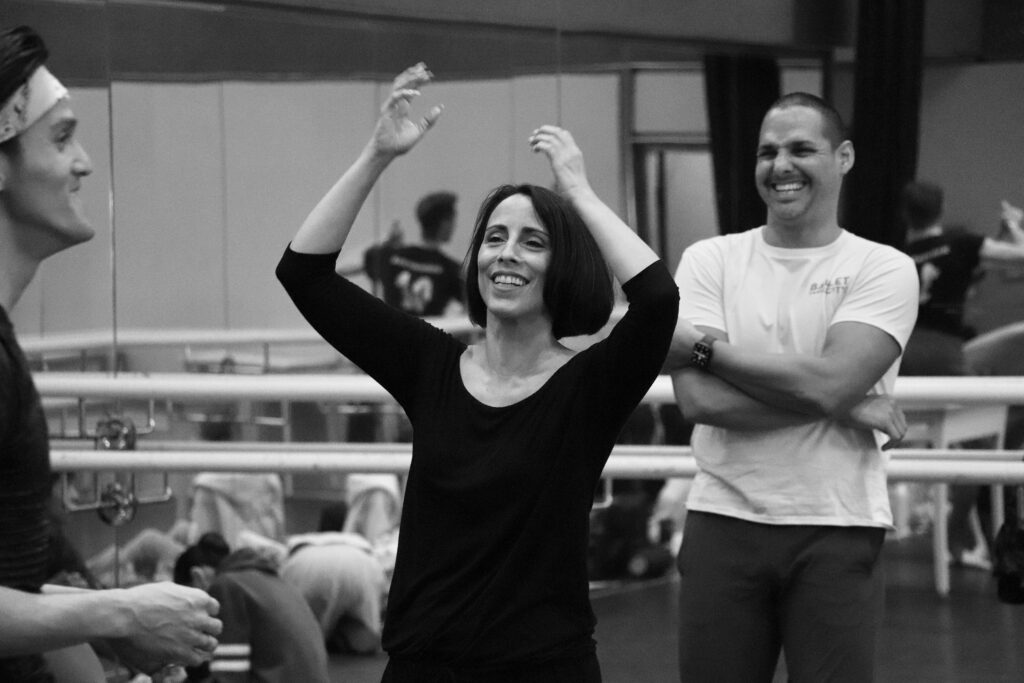 A black and white photo shows Annabelle Lopez Ochoa from the waist up as she raises her hands up, smiling, during a rehearsal. Two male dancers laugh with her in the background.