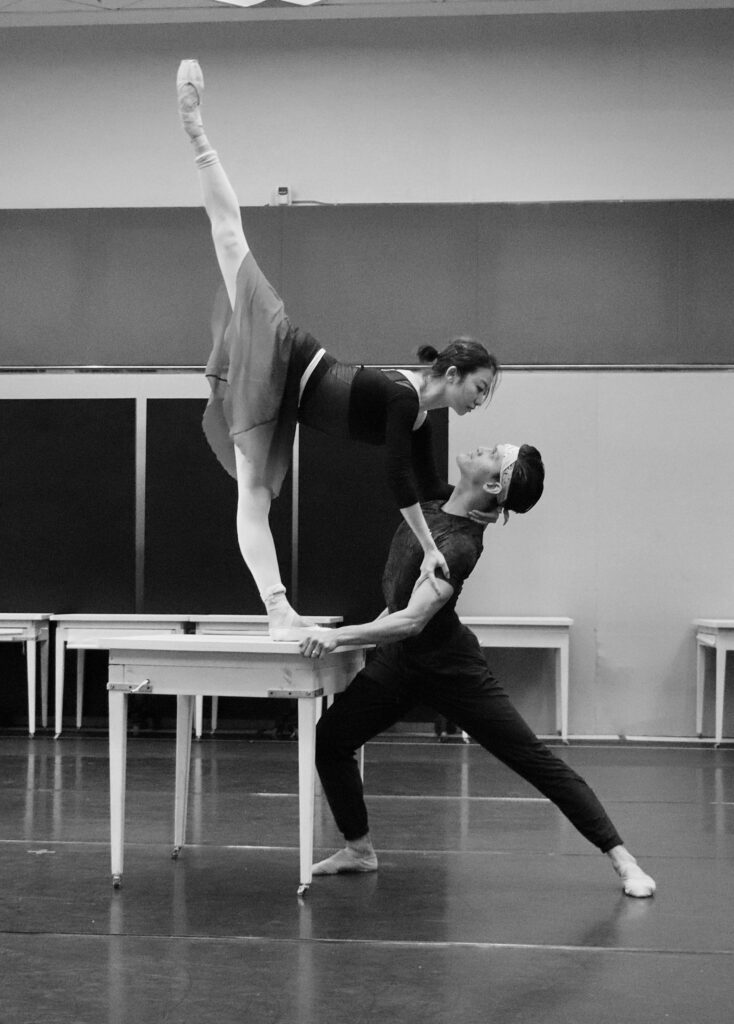 A black and white photo shows a male and a female dancer rehearsing together in a large studio with white roller desks behind them. The ballerina, wearing a dance sweater, tights, a leotard, a long practice skirt and pointe shoes, does an arabesque penché on one of the desks, holding onto her partner's shoulders. The male dancer, wearing a t-shirt, tights, ballet shoes and a bandana, lunges and holds her steady. They look into each others' eyes.