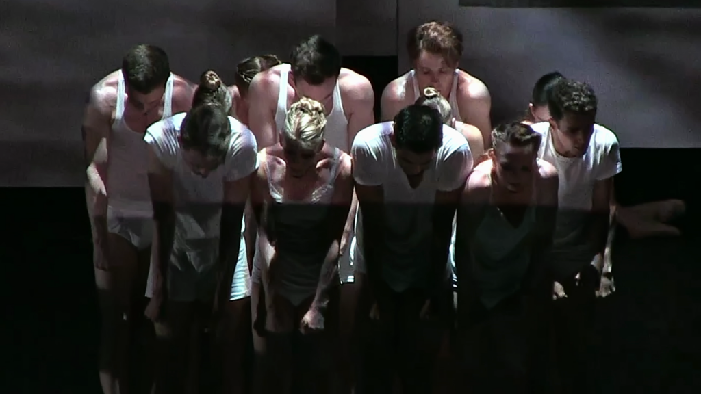 A still from a documentary shows a group of 11 male and female dancers huddled together, heads down, as they are lit from above. They all face the audience and clench their fists and shrug their shoulders up with tense emotion, dressed in minimalist white costumes.