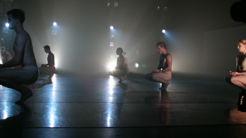 A still from a documentary shows a side view of a stage with five male and female dancers shown crouching to sit on their heels, hands laid gently on their laps. They wear minimalist neutral-colored costumes and look down solemnly.