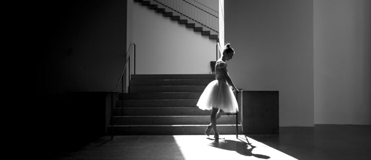 In this black and white photo, a balerina in a knee-length tutu stands at the bottom of a stairwell, holding onto the handrail and looking down at it. She stands in a pool of bright light under an archway, and in the background is another staircase perpendicular to the archway.
