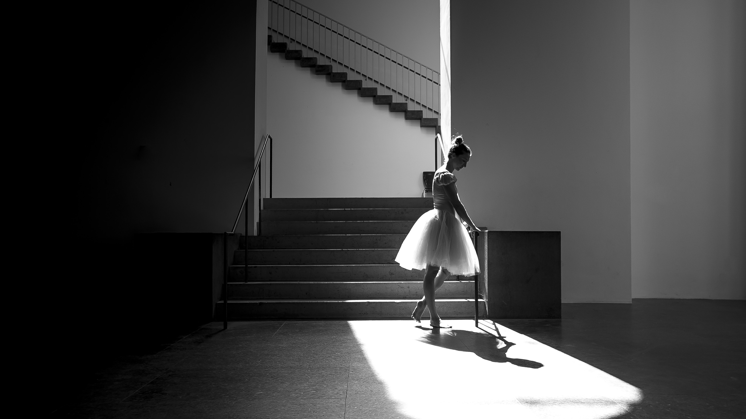 In this black and white photo, a balerina in a knee-length tutu stands at the bottom of a stairwell, holding onto the handrail and looking down at it. She stands in a pool of bright light under an archway, and in the background is another staircase perpendicular to the archway.