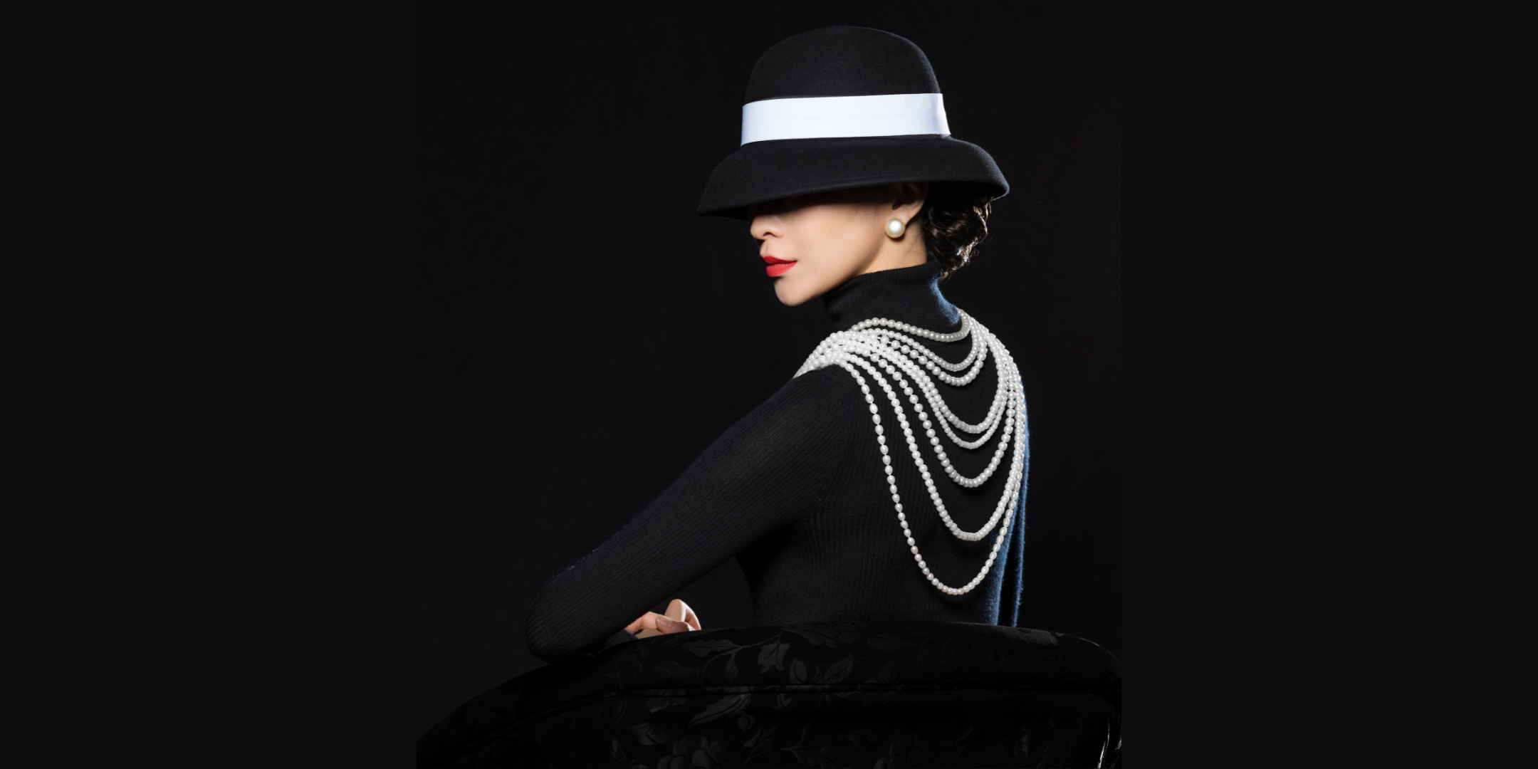 In front of a pitch black background, a female dancer in a sleek black turtleneck and fashionable hat (black, round top, with a white sash around the middle) that covers her eyes poses behind a black chair. She is shown chest-up and her body is turned sideways as she turns her chin toward the camera, a sly look on her face. She wears bright red lipstick, large pearl earrings and eight stacked pearl necklaces.