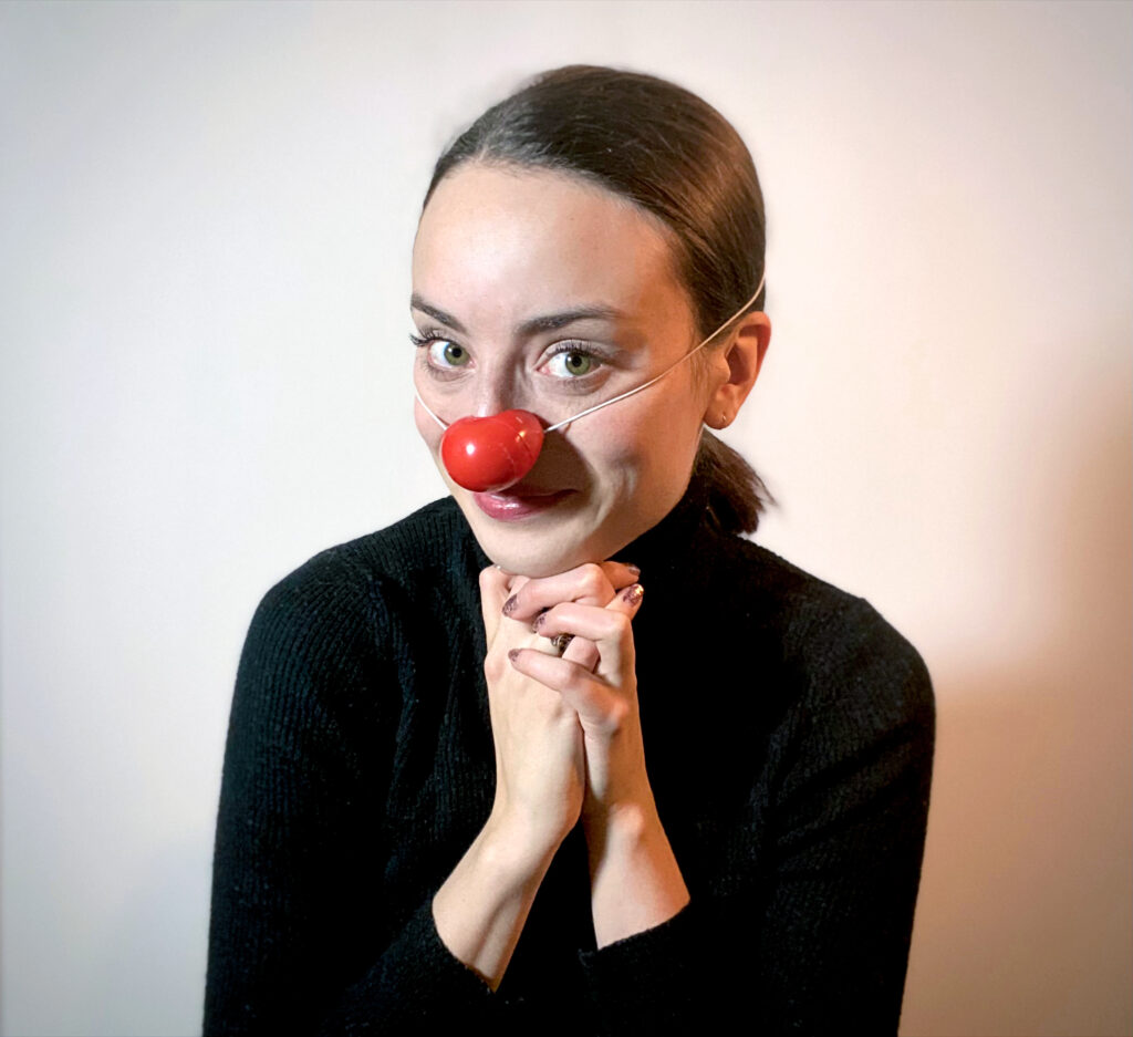 A headshot of Claire Davison wearing a red clown nose and black turtleneck, her hands clasped under her chin.
