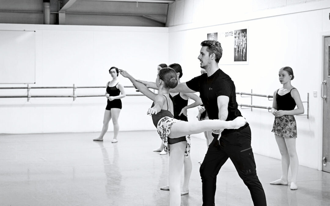 In this black and white photo, Iain Mackay, a ballet teacher, is shown working with a young female student as she poses in first arabesque. He stands behind her and uses his hands to lengthen her leg and front arm. Four other ballet students stand behind them and to their left, watching. The dancers wear pink tights, ballet slippers, leotards and ballet skirts. Mackay is wearing a black T-shirt and black athletic pants.