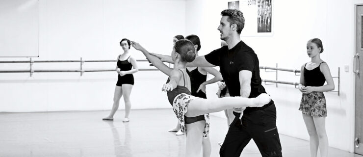 In this black and white photo, Iain Mackay, a ballet teacher, is shown working with a young female student as she poses in first arabesque. He stands behind her and uses his hands to lengthen her leg and front arm. Four other ballet students stand behind them and to their left, watching. The dancers wear pink tights, ballet slippers, leotards and ballet skirts. Mackay is wearing a black T-shirt and black athletic pants.