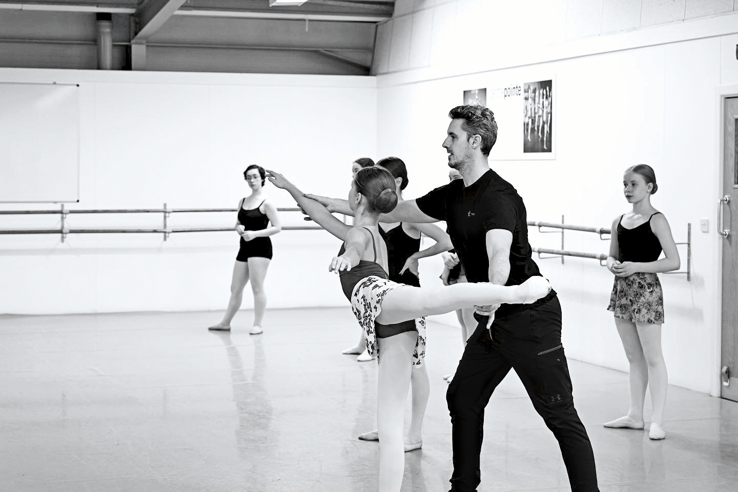 In this black and white photo, Iain Mackay, a ballet teacher, is shown working with a young female student as she poses in first arabesque. He stands behind her and uses his hands to lengthen her leg and front arm. Four other ballet students stand behind them and to their left, watching. The dancers wear pink tights, ballet slippers, leotards and ballet skirts. Mackay is wearing a black T-shirt and black athletic pants.