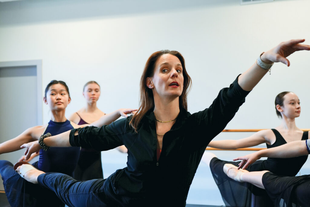 Jill Krutzkamp is shown from the waist up demonstrating a first arabesque, looking out into the mirror in front of her as she talks to a group of dancers behind her. The dancers mimic her position and wear leotards, tights and ballet skirts. Krutzkamp wears a black zip-up shirt, dark jeans and open-toed sandals.