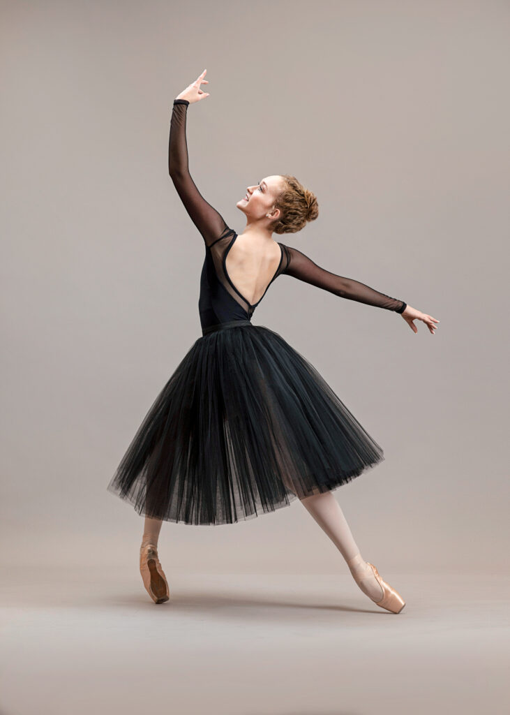 Kayla DeGaray poses in front of a tan backdrop, wearing a long black practice tutu and long-sleeved balck leotard with mesh sleeves. Shown in profile, she poses on pointe in an effacé lunge with her right leg in front and right knee bent. She twists her upper body, looking up at her raised left arm and stretching her right arm behind her.