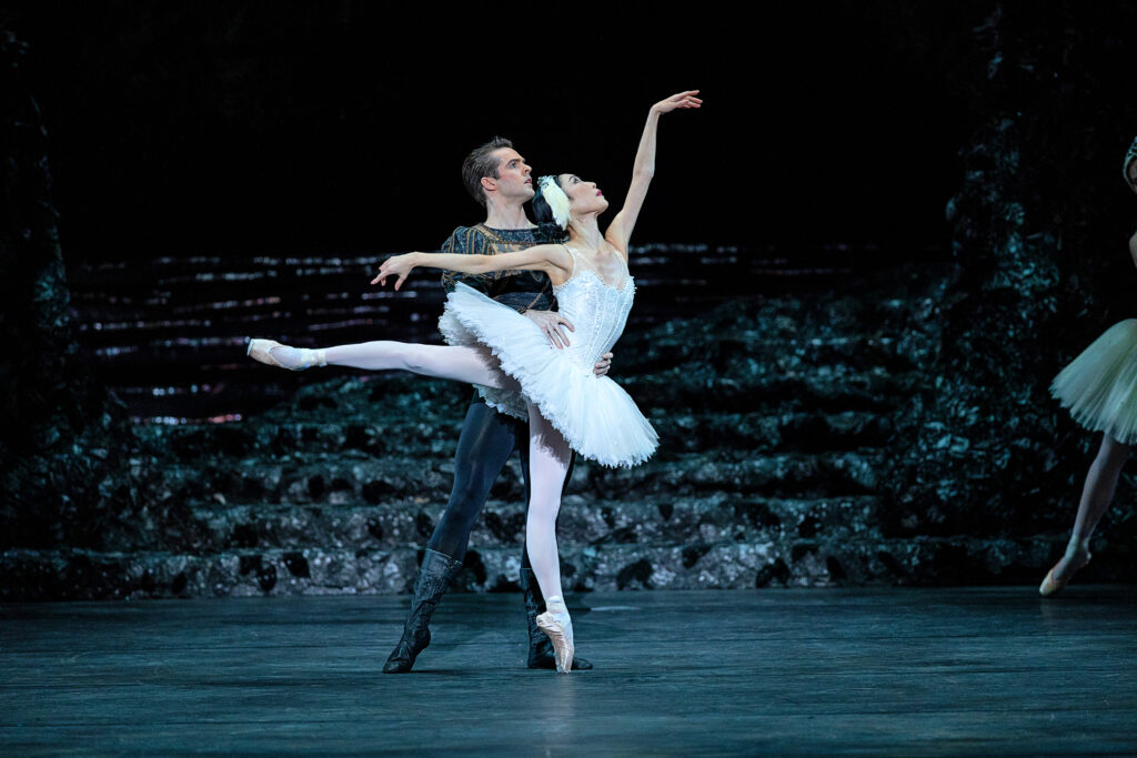 Momoka Hirata and Lachlan Monaghan dance a pas de deux from Swan Lake onstage during a performance. Hirata stands on her left foot on pointe in first arabesque as Monaghan stands behind her, holding her waist with both hands and and reaching his right leg out in tendu. She wears a white tutu and feathered crown, pink tights and pointe shoes; he wears black tights and leather dance boots, and a dark tunic with puffed upper sleeves.