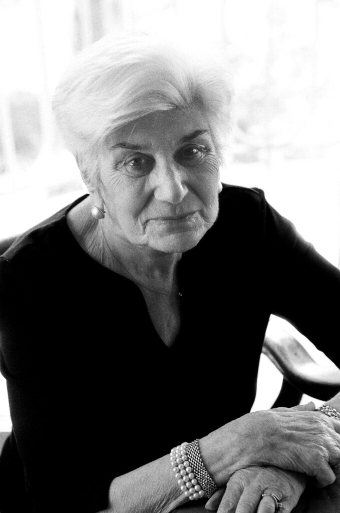 A black and white photo shows Naomi Warren, an elderly woman with short white hair wearing a black blouse, waist-up. She rests her hands on a table gently, leaning in toward the camera with a slight smile.