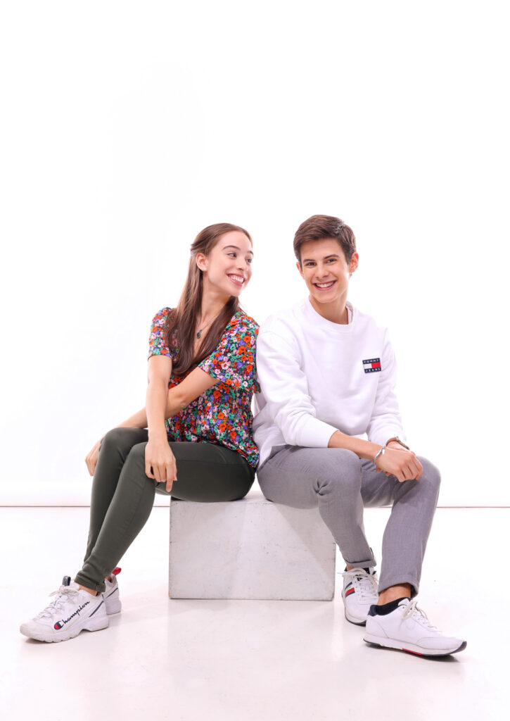 António Casalinho and Margarita Fernandes sit side-by-side on a white box in front of a white backdrop. They lean against each other affectionately, smiling broadly. Casalinho wears a white sweatshirt, gray weatpants and white sneakers, while Fernandes wears a floral-patterned short-sleeved top, olive green leggings and white sneakers.