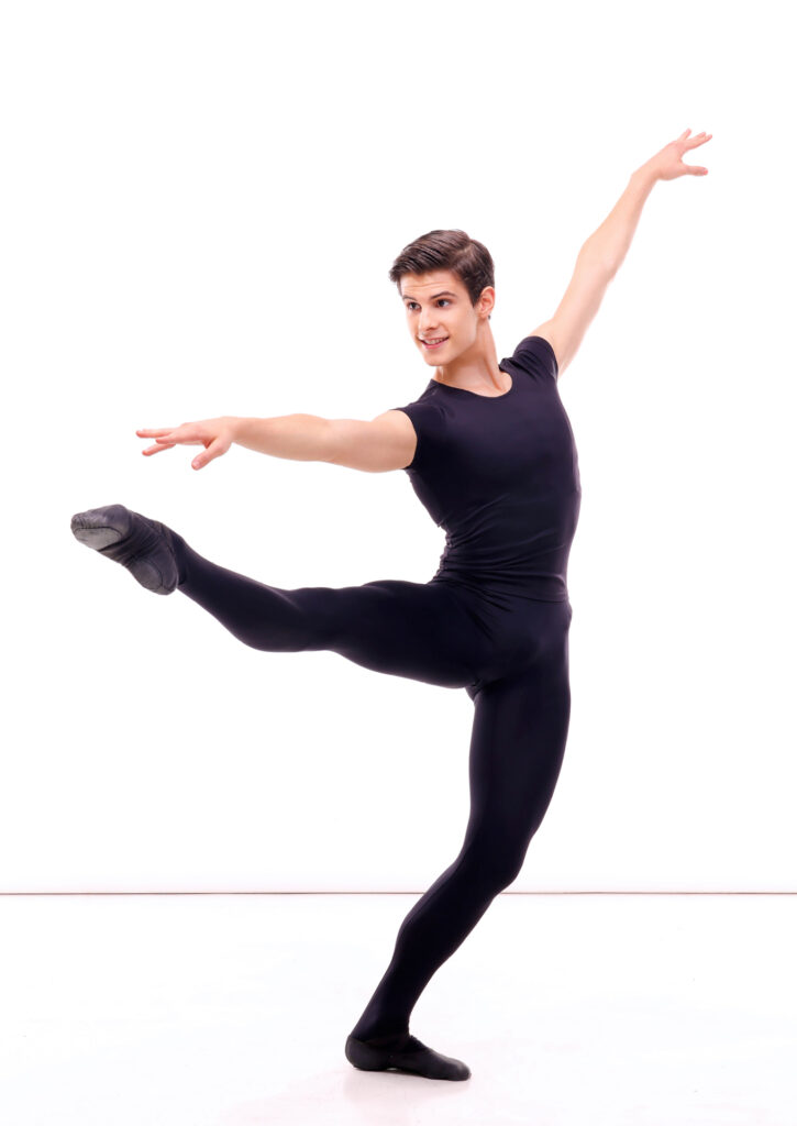 Antonio Casalinho, wearing a black T-shirt, tights and ballet slippers, stands in ecarté with his left (standing) leg in plié and his right leg at 90 degrees. He lifts his left arm up and extends his right arm out over his leg, and looks in the direction of his leg with an enthusiastic smile.