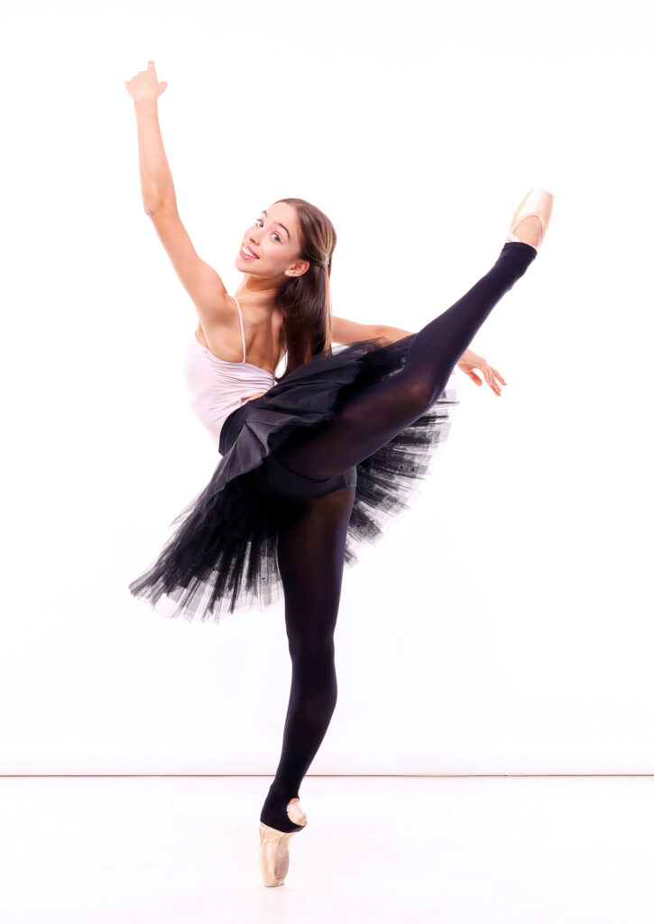 Margarita Fernandes poses in front of a white backdrop and wears a pink leotard, black tights, a black practive tutu and pink pointe shoes. She faces the upstage right corner, stands on pointe her right leg and does a high attitude derriere with her left leg. She holds her left arm high and looks back over her left should, smiling brightly with a large smile toward the camera. Her long brown hair falls down her back.